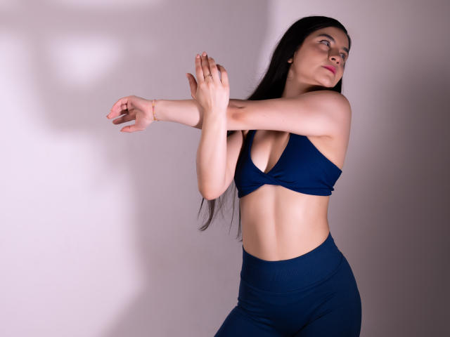 Jade Harpper, avec ses longs cheveux noirs, son débardeur de sport bleu et son legging assorti taille haute, les bras levés, montrant sa petite poitrine et son ventre musclé, dans une lumière douce..