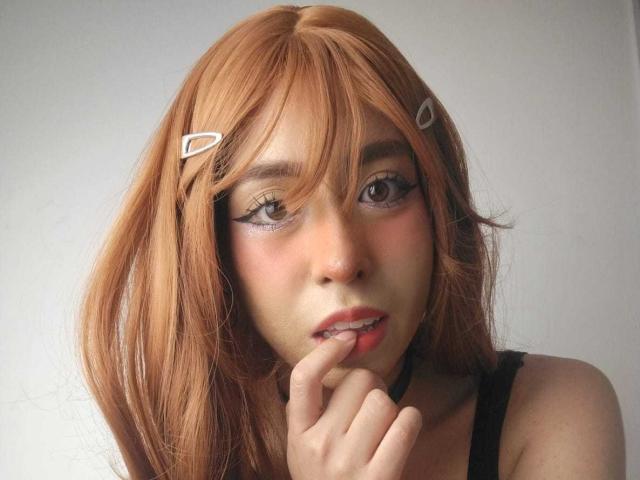 Amelie Benson con una parrucca arancione, mollette per capelli bianche, ombretto rosa e un dito vicino alle sue labbra rosa. Sembra carina, con una mano che tocca il mento. Indossa una maglietta nera..