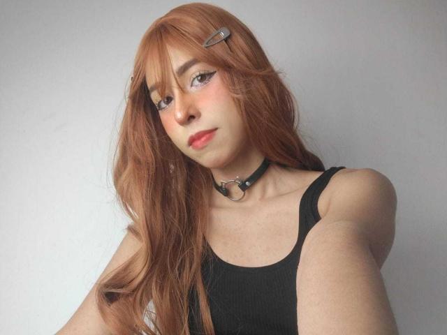 Amelie Benson tem cabelos longos, avermelhados, um colar preto com um pingente, e veste uma regata preta, olhando para cima com batom rosa e maquiagem discreta..