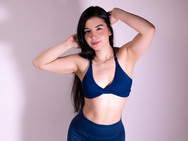 Jade Harpper, avec ses longs cheveux noirs, son haut de bain bleu à encolure américaine et sa jupe assortie, les bras levés, souriante, montrant ses abdominaux tonifiés et ses petites seins, se tient devant un fond uni..