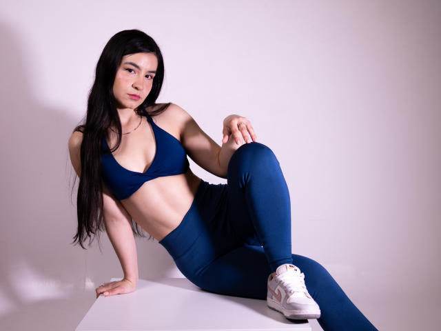 Jade Harpper, met lang zwart haar, een blauwe sportbeha en bijpassende broek, zittend op een wit oppervlak, waarbij haar borsten en billen zichtbaar zijn, draagt witte sneakers..