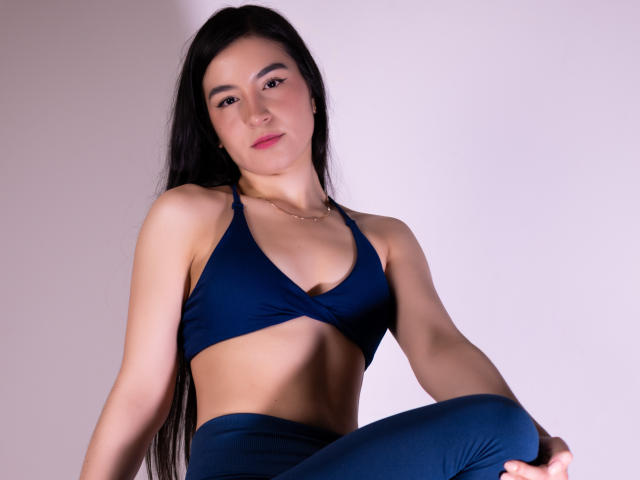 JadeHarpper with long black hair, wearing a navy blue sports bra and matching pants, sitting with a confident smile, showing her small tits and slim, toned abs.