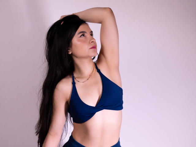 JadeHarpper with long black hair, wearing a blue bikini top, arm raised, looking to the side, small gold necklace, pale pink background, slightly visible abs, confident expression.