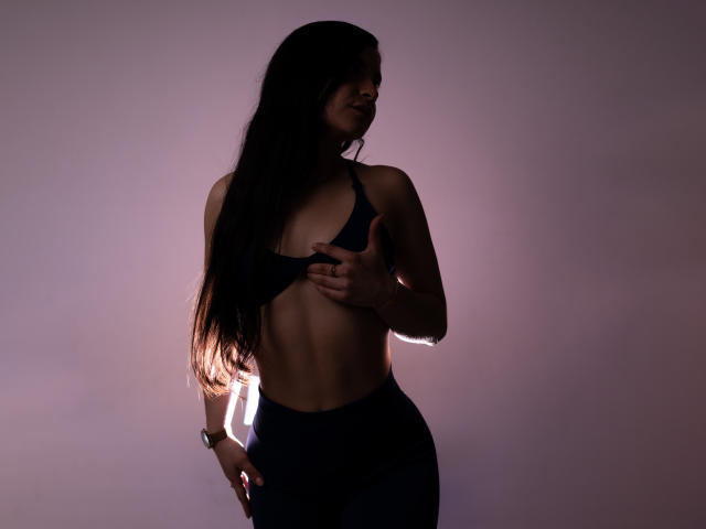 JadeHarpper with long dark hair, wearing a black sports bra and high-waisted pants, covering her small tits with one hand, standing in dim light, shadow on her face.