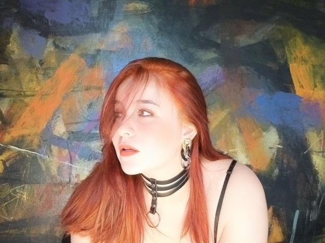 AngelaMonticelli with red hair, wearing a black choker with a ring, looking to the side, has earrings, and a black top, her tits slightly visible, in a colorful background.