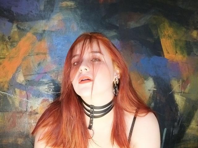 AngelaMonticelli with red hair, black strappy collar, and earrings, looking up with slightly parted lips. She's a pale-skinned, attractive chick with a sexy, goth vibe.