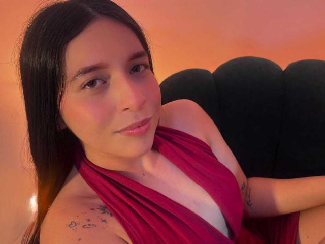 ValentinaRiva with long black hair, red halter dress, and tattoos on her right arm, sitting on a black cushion, smiling softly, in a warm orange-lit setting.