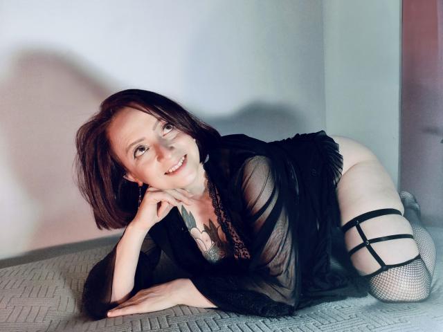 CatalinaMonyque, con i capelli castani corti, biancheria intima nera trasparente e collant a rete, è a quattro zampe, sorridente, mostrando i suoi seni e il sedere scoperto, su un letto con una superficie strutturata..