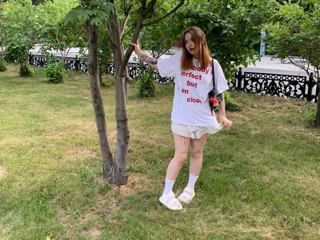 MaritaPavelko with pale skin, red hair, white tee, white skirt, white socks, yellow sneakers, standing by a tree, grassy area, black ornate fence, red bracelet, smiling.