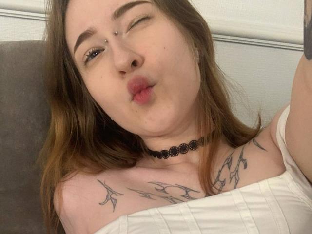 MaritaPavelko with brown hair, winking, puckering lips, white top, black choker, tattoos on shoulders, nose piercing, light skin, and a cute, relaxed expression.