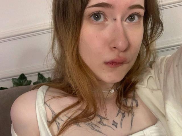 MaritaPavelko with pale skin, brown hair, blue eyes, nose piercing, lip piercing, white off-shoulder top, chest tattoos, choker, looking at camera, slightly parted lips, soft expression.