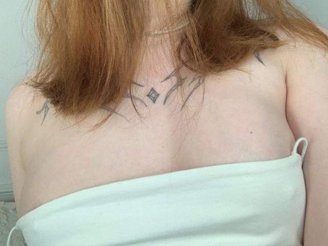 MaritaPavelko's bare tits, covered by a white strapless top, show small black star tattoos on her chest. Her red hair is slightly messy.