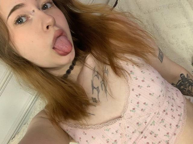MaritaPavelko with tongue out, pink floral top, tattoos, nose piercing, choker, and blonde hair. Her tits are visible, and she's looking at the camera with blue eyes.
