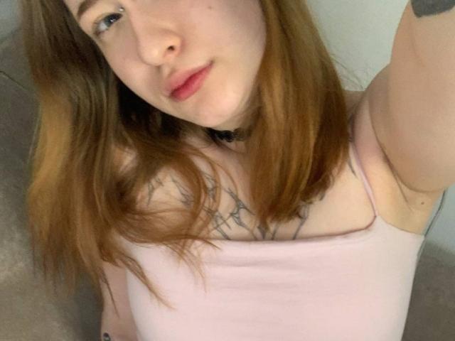 MaritaPavelko with light brown hair, pink tank top, small tits, tattoo on chest, choker necklace, smiling, arm raised, selfie, fair skin, light pink lips.
