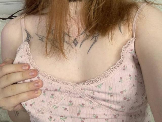 MaritaPavelko's chest with small tits, lace-trimmed pink tank top, red tattoo on upper chest, hand covering left tit, pale skin, red hair.