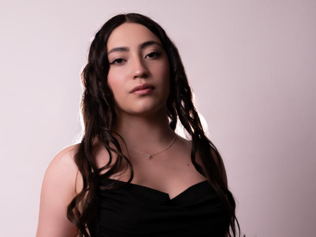 AnnFisher has long, wavy brown hair, a light skin tone, and is wearing a black strapless top, with a small necklace, looking directly at the camera with a neutral expression.