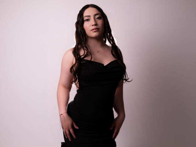 AnnFisher with long, wavy brown hair wearing a black, strapless dress, hands on her black skirt, standing, looking at the camera, small necklace, slight smile, soft light.