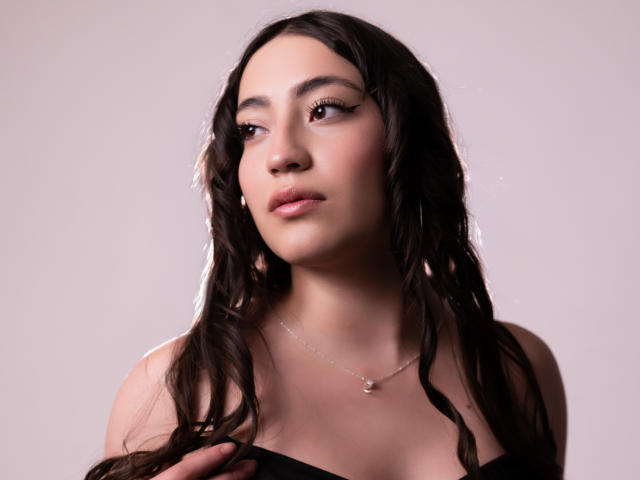 AnnFisher has wavy black hair, light brown skin, and a small gold necklace. She's looking to the side with a neutral expression, wearing a black strap top.