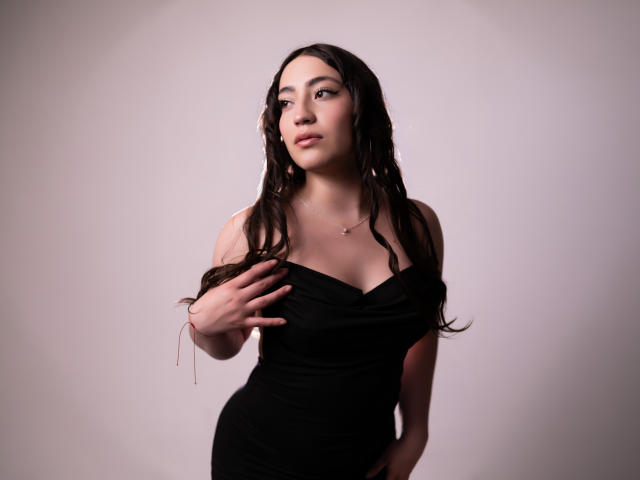 AnnFisher with long dark dreadlocks, wearing a black off-shoulder dress, touching her chest, showing her tits, looking to the side, pale skin, subtle makeup, necklace, red bracelet.