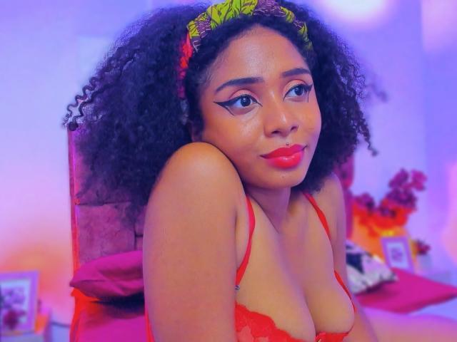 MeganWilliams with curly black hair, red lipstick, and red lingerie, sitting on a bed, showing her tits, wearing a green headband, smiling, with blue and pink lighting.