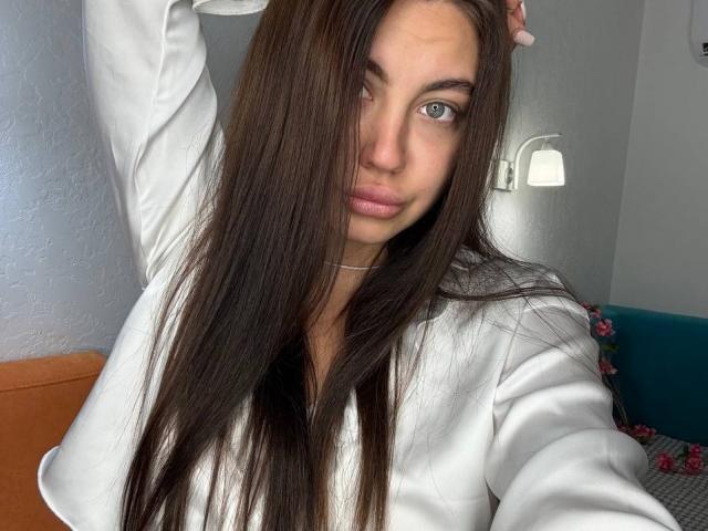 InesGueningsman with long brown hair, blue eyes, white shirt, arm raised, looking at camera, subtle pink lips, small silver necklace, soft natural light, casual selfie.