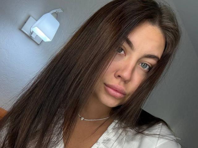 InesGueningsman with long brown hair, light skin, blue eyes, and pink lips, wearing a white shirt and a silver necklace, looking into the camera with a neutral expression.