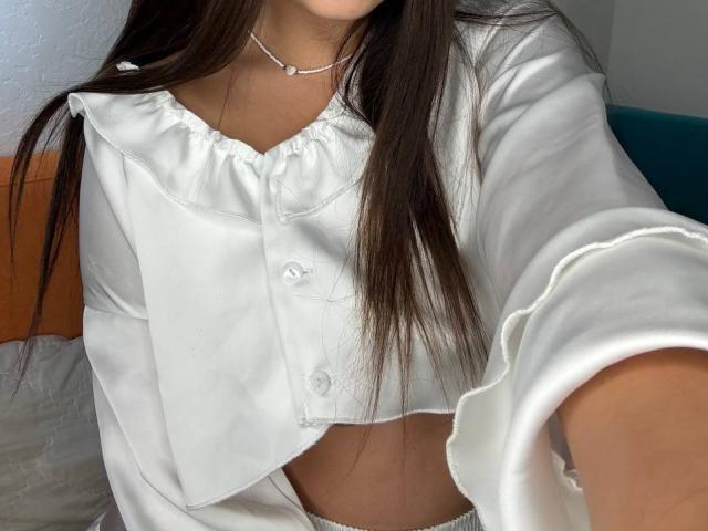 A camisa branca de botões de Ines Gueningsman, que mostra sua barriga, com cabelos castanhos longos, usando um colar, estendendo o braço em direção à câmera. Seios não visíveis..