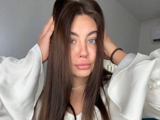 InesGueningsman with long brown hair, hands in hair, wearing a white shirt, blue eyes, light skin, subtle makeup, slight pout, thin necklace, air conditioning unit in background.
