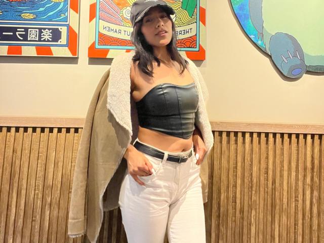 BrianaHarpers with taut tits in a black strapless top, white pants, beige coat, and gray cap, standing with hands in pockets, wooden wall in background.