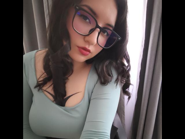 Susan Monroy, met krullend donker haar, een bril en een lichtgroen top, laat haar borsten zien in een top met een lage halslijn, rode lippen en een serieuze uitdrukking..
