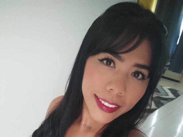 NinaPalmerX with black hair, brown eyes, and pink lipstick, smiling at the camera. She's got a tattoo just below her chin. Her tits are partially visible.