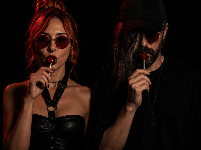 ModeeviL with red hair, heart-shaped sunglasses, and leather top, sucking a red lollipop. ModeeviL with long hair, black cap, and beard, sucking a red lollipop.