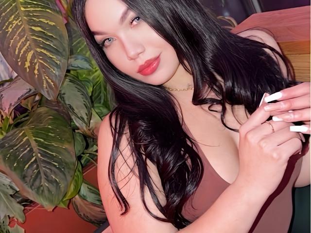 MiaDenver with long black hair, red lipstick, and a tan bra, showing her white-manicured nails, holding her cleavage, and smiling gently. Her tits are partially visible.