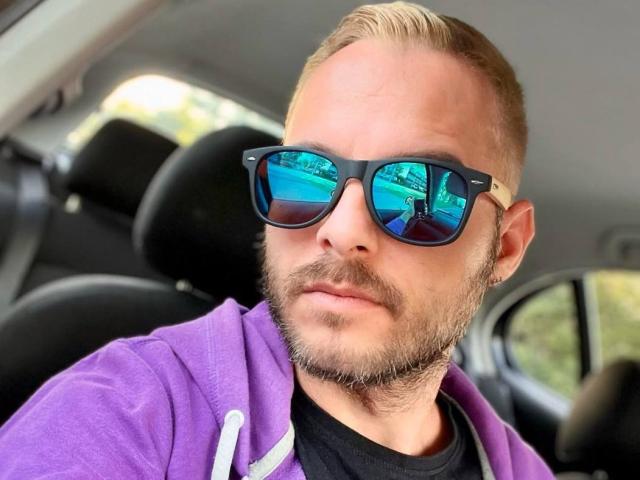DamienJoy, avec une barbe blonde, portant des lunettes de soleil noires reflétant un ciel bleu et des arbres, un sweat à capuche violet et un t-shirt noir, assis dans une voiture..