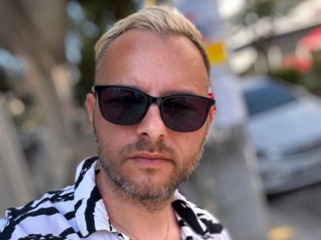 DamienJoy with blond hair, beard, and sunglasses, wearing a black and white striped shirt, looking serious. Boob-free, ass-free, pussy-free. Blurry background, car, and person in the back.