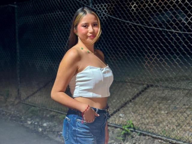 AmilyRizzo with tan skin, brown eyes, and long blonde hair in a white strapless top, showing her small tits, and blue jeans, standing by a chain-link fence.