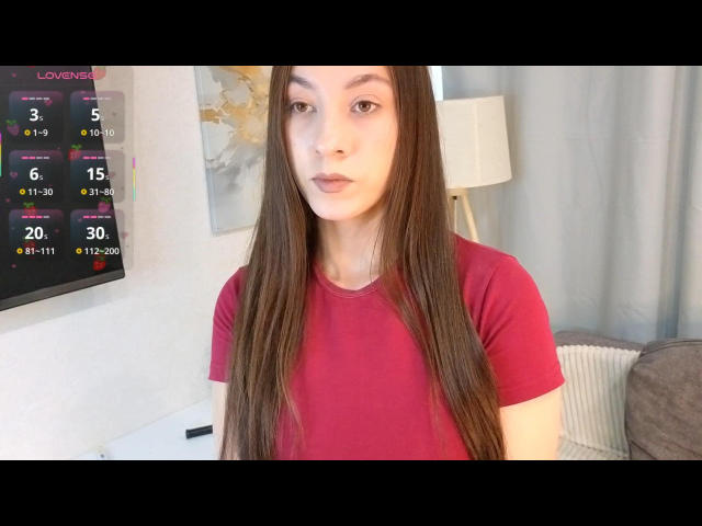 AngelStusy, con lunghi capelli castani, una maglietta rossa e un'espressione neutra, è in piedi in una stanza con una parete bianca e una tenda grigia. Ha un viso esile e piccoli seni..