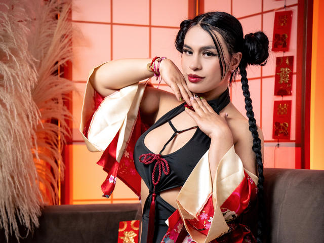 GreizSubKimura con moños negros dobles, vistiendo un kimono rojo y dorado, un top halter negro que muestra el escote, una cinta roja y pulseras, con las manos cerca de la cara, sentada en un sofá marrón..