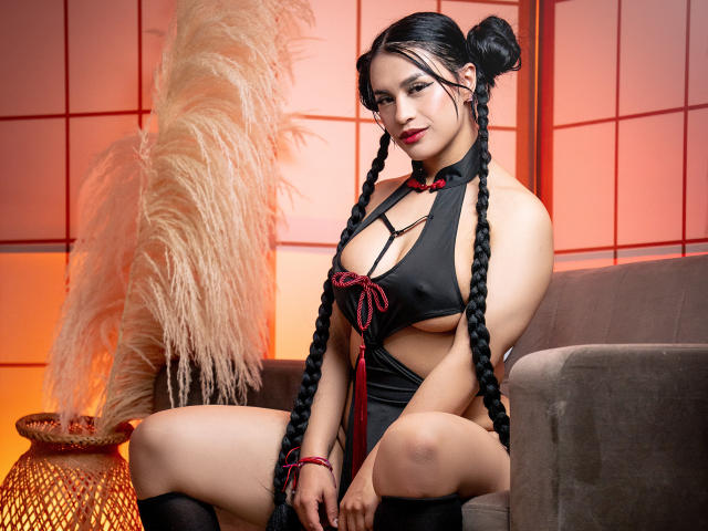 GreizSubKimura, con due trecce nere, indossa un vestito nero scollato che mette in mostra il suo seno e i piccoli fiocchi rossi, ed è seduta su un divano grigio, mostrando il suo sedere..
