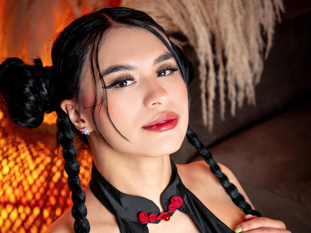 GreizSubKimura con trecce nere, rossetto rosso e trucco lucido, indossa un top nero con spalline e bottoni rossi, guarda verso l'alto con un leggero sorriso, con una luce calda sullo sfondo..