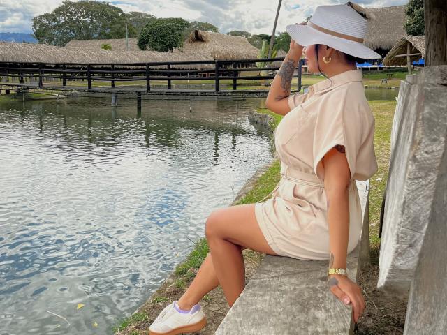 JessicaFiore with tan skin and tattooed arm, wearing a beige dress and sun hat, sitting on a stone edge by a waterway, white sneakers, holding hat, looking away.