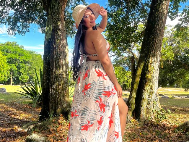 CatalinaRivero with long blue hair, tan skin, and a cute ass, wearing a floral dress and hat, looking back with a smile, standing between trees in a sunny park.