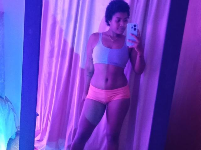 Caylin with short curly hair, wearing a gray sports bra and pink shorts, holding a phone, standing in front of a mirror, showing a toned, tattooed, and curvy body.