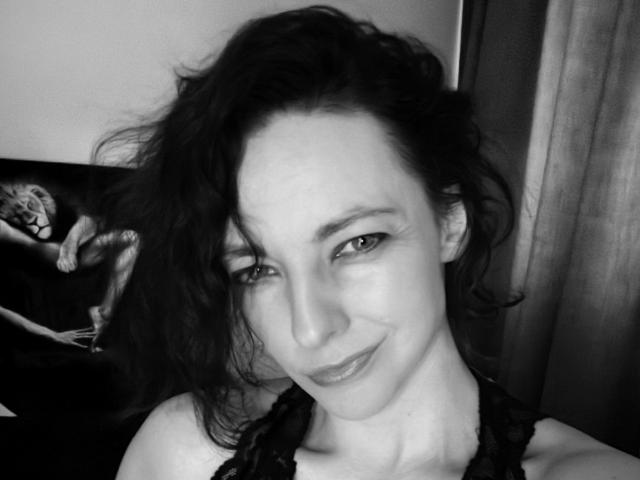 BlackParty, con cabello oscuro y ondulado, sonriendo sutilmente, vestida con una blusa negra de encaje, mostrando una nariz delicada y ojos relajados, en un selfie en escala de grises. Sus senos no son visibles..