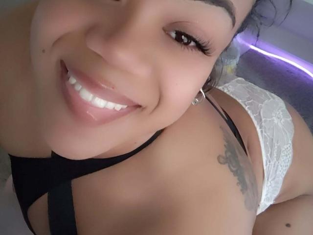 LotusFineAss sorrindo com dentes brancos, vestindo um top preto com tiras e calcinha de renda branca, mostrando uma tatuagem no ombro direito, olhos castanhos e cílios longos, com brincos de argola..
