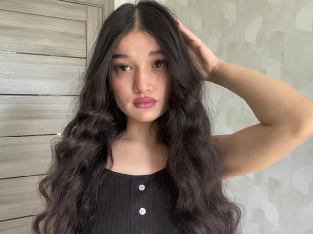YoulandaCarlough with long, wavy black hair, wearing a black ribbed top with buttons, has her hand in her hair, looking serious with pink lipstick.