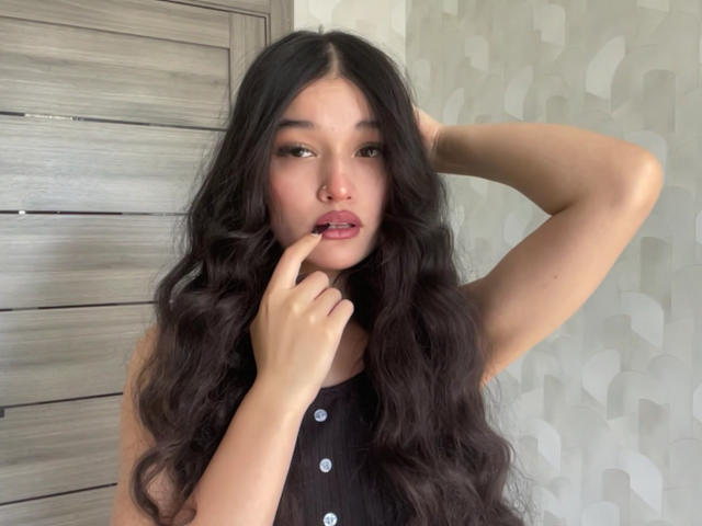 YoulandaCarlough with long wavy black hair, brown eyes, and pink lips, touching her finger to her mouth, wearing a black sleeveless top with white buttons.