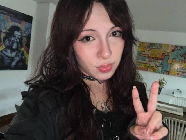 ValeryBeaumont with long dark hair, black choker, and black top, making a peace sign with pink lipstick, tattoo on chest, and slight pout, in a casual selfie.