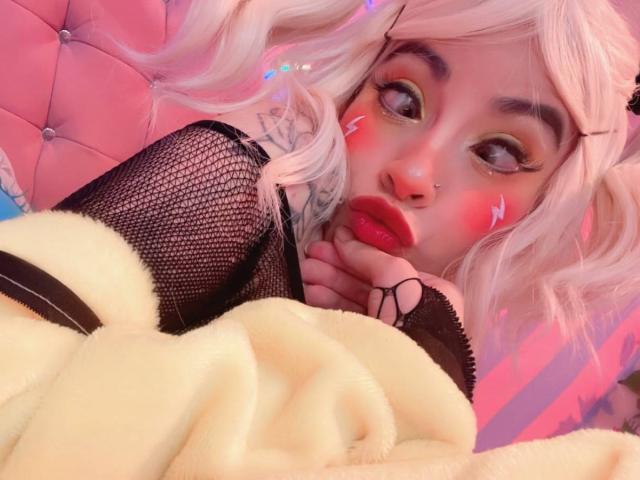 LaurenEvie69 with pink pigtails, red lipstick, and fishnet sleeves, lying on a pink tufted bed, wearing a yellow blanket, and sporting a nose ring.