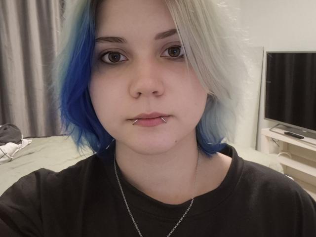 PaolaStohler with blue and white hair, nose ring, lip piercing, black shirt, and silver necklace, staring at camera. Her eyebrows are thick, and her skin is pale.
